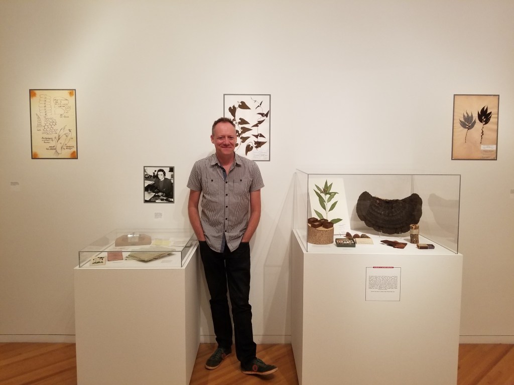 Dr. Eric Tepe standing next to a herbarium exhibit featuring specimens from UC's herbarium